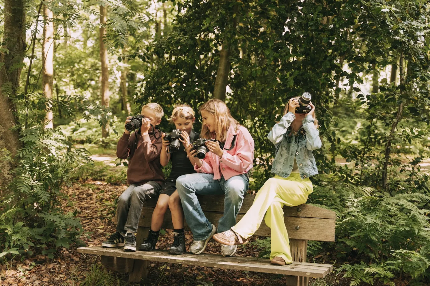 Vier kinderen op een houten bankje in het bos fotograferen tegelijk verschillende kanten op met hun spiegelreflexcamera's