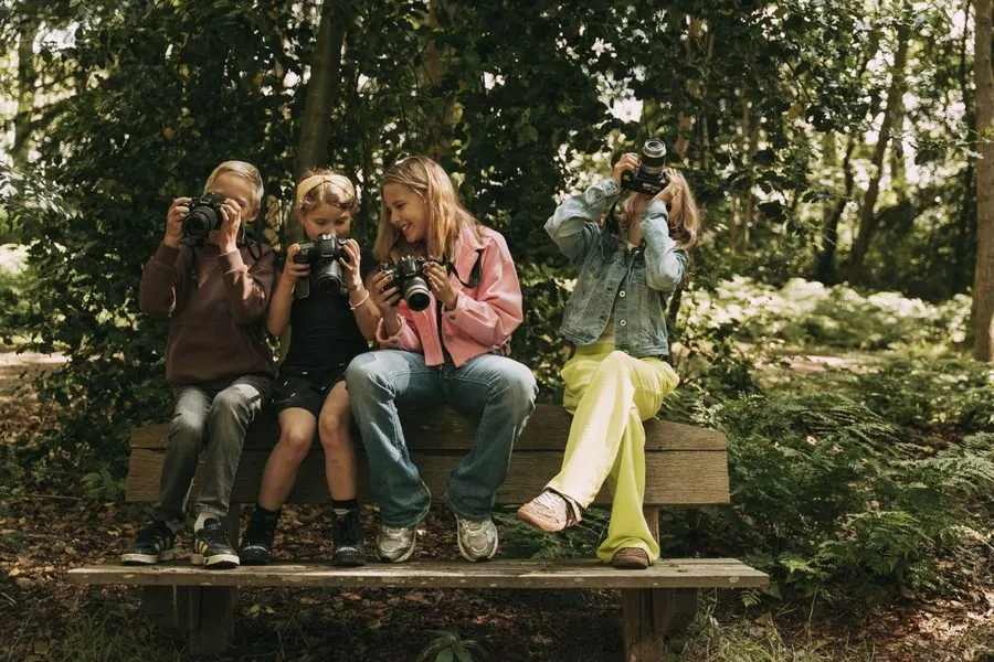 Groepje kinderen zit lachend naast elkaar op een houten bankje in het bos, elk met een spiegelreflexcamera