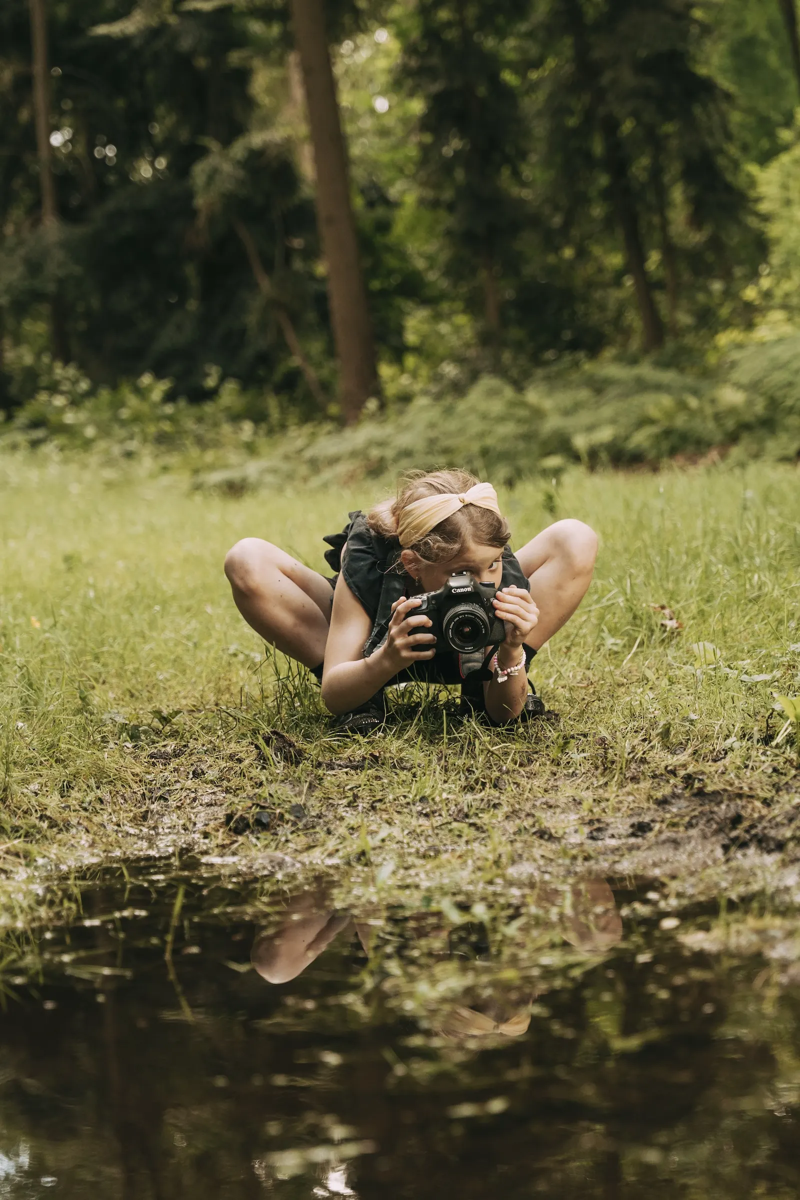 Meisje ligt laag bij de grond in het gras en fotografeert met een spiegelreflexcamera, met haar spiegelbeeld in het water