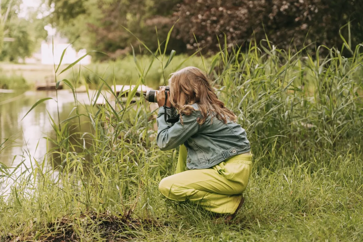 Meisje hurkt tussen het riet aan de rand van een vijver en fotografeert iets in het water met haar spiegelreflexcamera